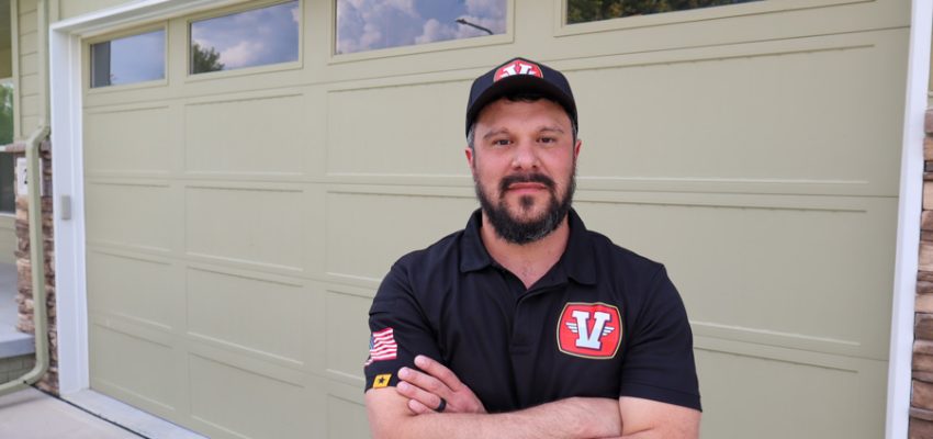 A professional Garage Door Vets technician stands with arms crossed in front of a beige residential garage door, showcasing the brand's uniform and logo.