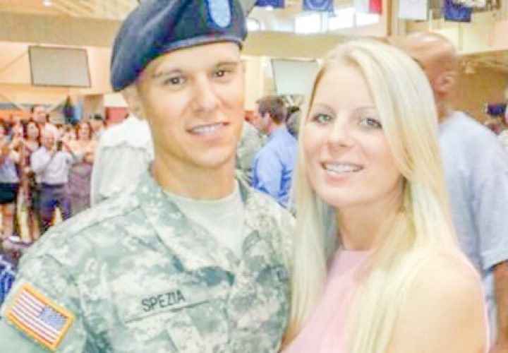 A young U.S. Army soldier in full uniform stands beside a woman in a pink dress at an indoor military ceremony, with flags hanging overhead and a crowd in the background.