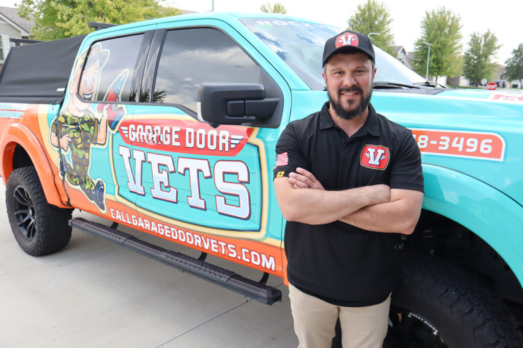 The owner of Garage Door Vets stands with arms crossed beside his brightly wrapped turquoise service truck, smiling at the camera to convey trust and professionalism for local garage door repair services.